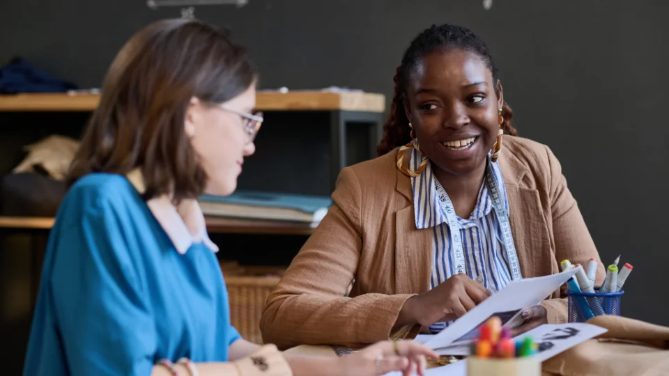 En lærer og en elev sidder over for hinanden og fører en samtale, mens de sidder ved et bord i et klasseværelse omgivet af studiematerialer og farvetuscher.