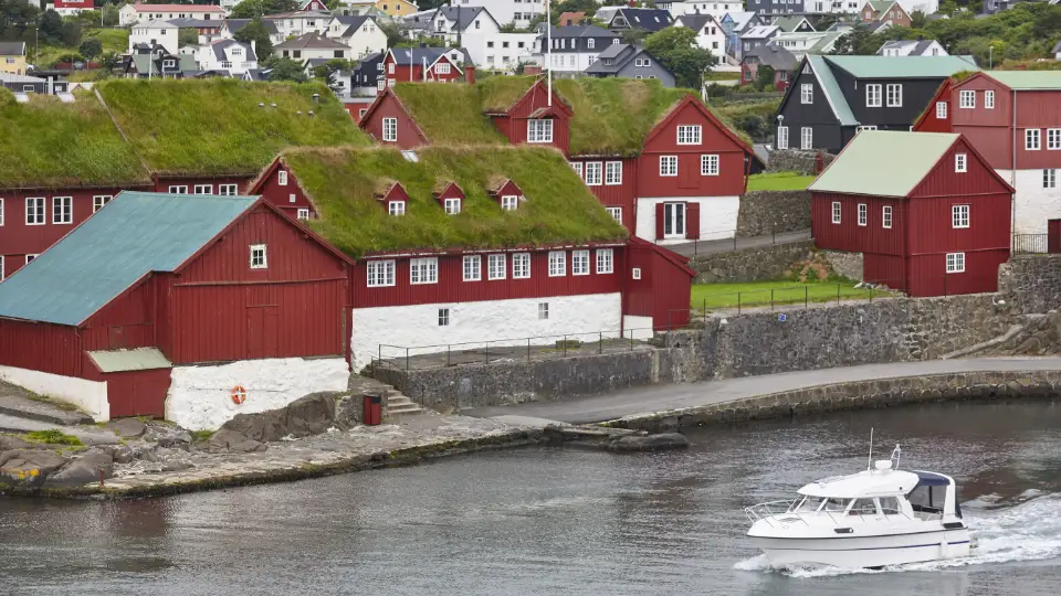 Et billede af traditionelle røde færøske huse med græstag ved en havnefront med en motorbåd, der sejler forbi.