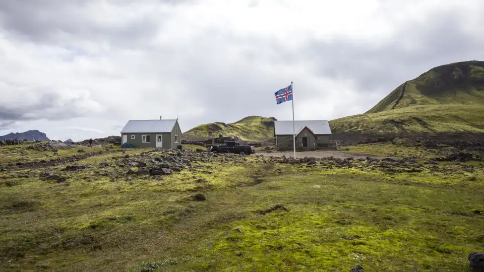 A rural scene in Iceland. There are two small buildings, both with white roofs, situated on a grassy and rocky landscape with hills in the background. In front of the building on the right, there is a flagpole with the Icelandic flag flying. A black vehicle is parked between the buildings. The sky is overcast with clouds, adding to the rugged and remote atmosphere of the location.