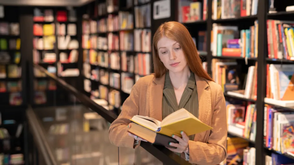 En ung kvinne leser en bok i en bokhandel med mange bøker i bakgrunnen.