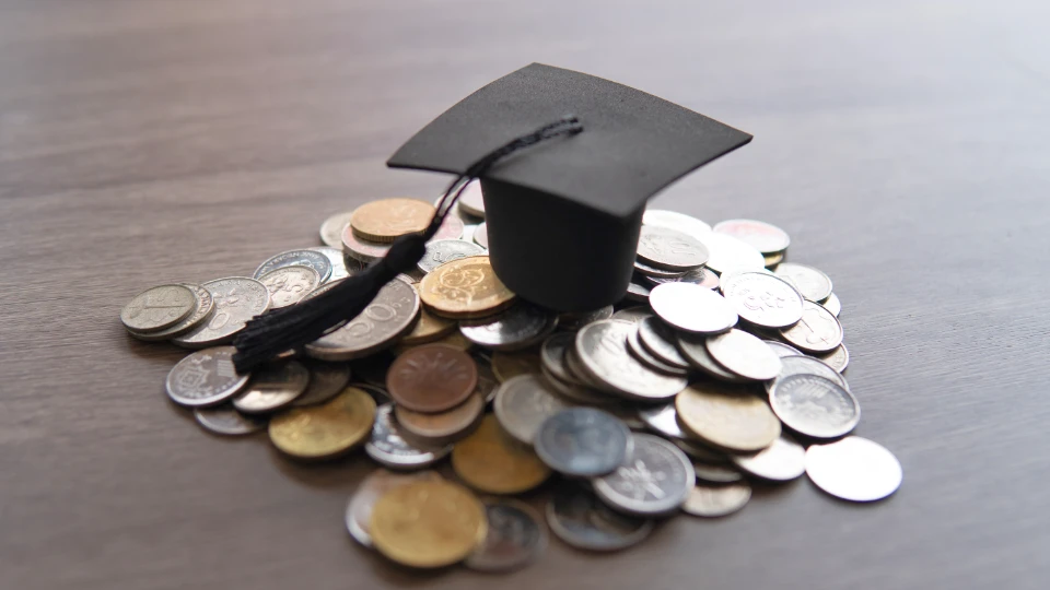 Bildet viser en svart akademisk hatt (mortarboard) plassert oppå en haug med mynter av forskjellige valutaer på et trebord. Dette kan symbolisere kostnadene ved utdanning eller investeringen i en akademisk fremtid.