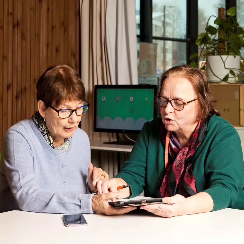 To ældre kvinder sidder ved et bord og bruger en tablet, mens de koncentreret diskuterer