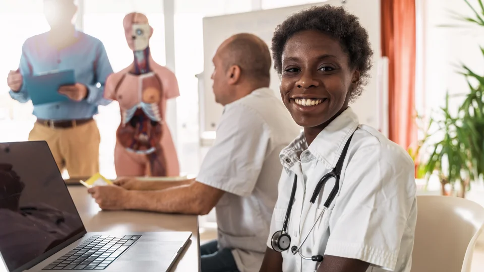 Woman in medical clothes smiling in a conference room with medical equipment in the background.