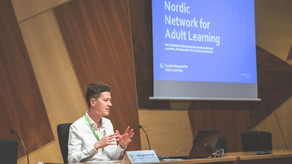 Male speaker presenting at a table with a "Nordic Network for Adult Learning" presentation projected on a screen in the background.