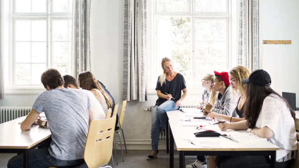 En gruppe studerende sidder i et klasselokale og arbejder ved borde, mens en kvindelig lærer sidder på kanten af et bord og taler med dem.