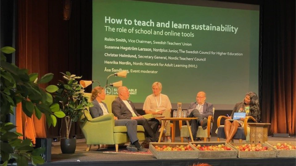 Panel discussion on sustainable education with speakers from Nordic educational organizations, seated on stage with green decor and baskets of apples, under a screen displaying the topic "How to teach and learn sustainability: The role of school and online tools."