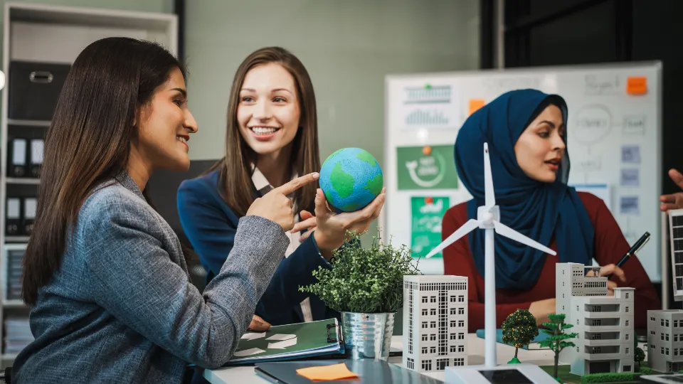 Tre kvinder i et mødelokale arbejder med en model af en vindmølle og miniaturebygninger; en kvinde holder en lille globus og smiler, mens en anden bærer hijab og skriver noter.