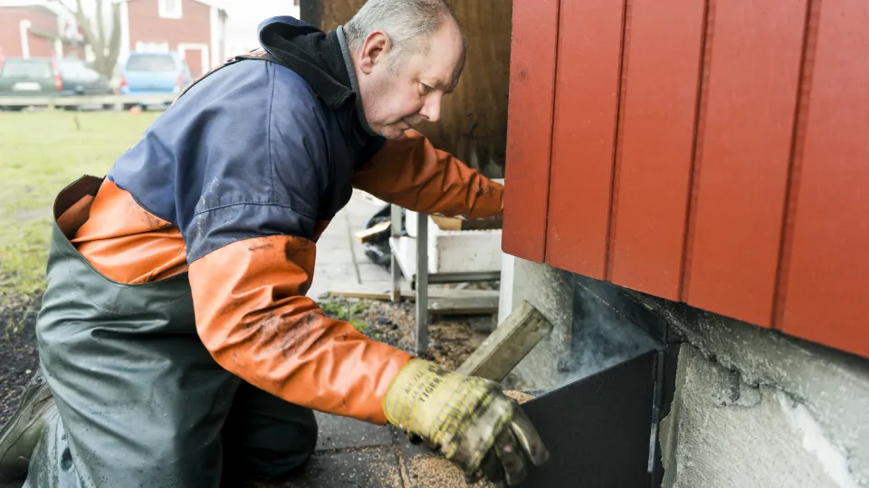 En mann iført arbeidstøy jobber med fundamentet på en rød bygning. Han bruker verktøy og ser konsentrert ut.