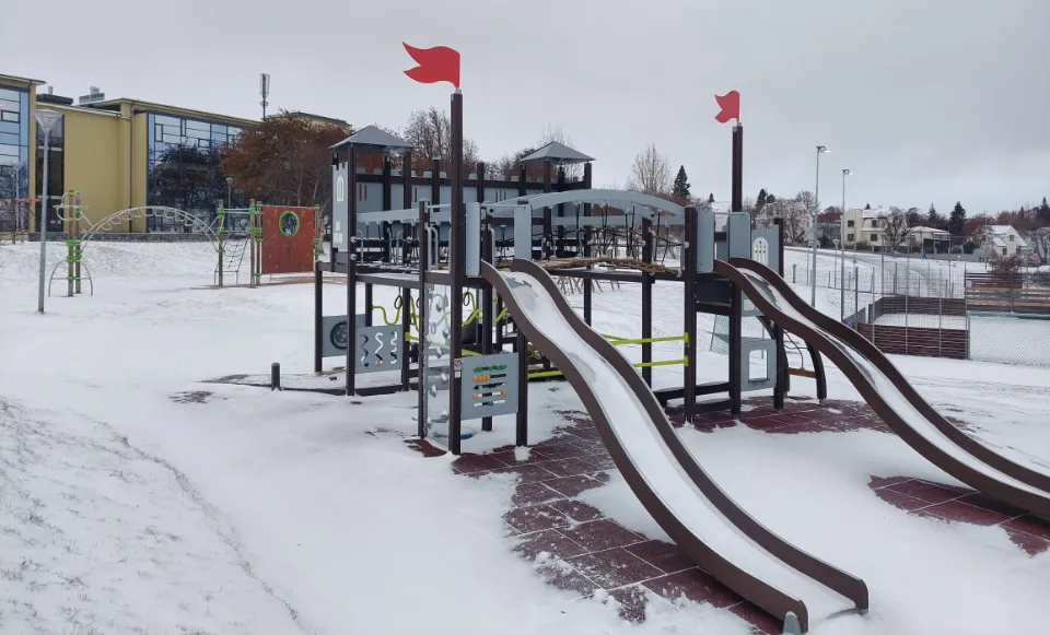 En snødekt lekeplass utenfor Brekkuskóli i Akureyri. Lekeplassen har flere rutsjebaner, klatrestativer og små flagg på toppen av strukturen. Området rundt har snø og noen bygninger i bakgrunnen.